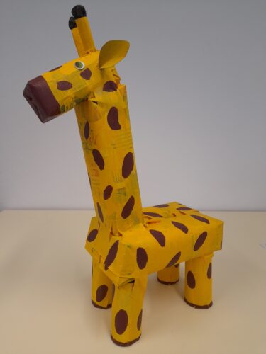 Personagem principal da obra - a girafa Olímpia<br/>A girafa foi elaborada com embalagens da Compal, de tamanhos diferentes. Sendo as pernas em rolos de papel de cozinha. Após a montagem, com apoio de fita e cola quente, foi pintada com guaches.